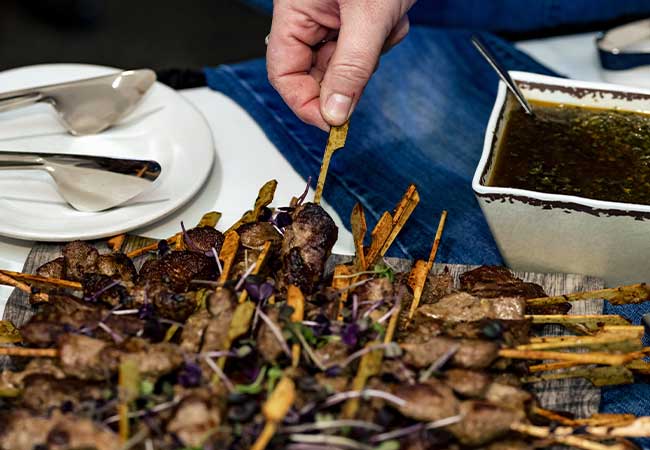 A hand picks up a skewer of grilled meat at a Complete Catering platter beside a dish of sauce.